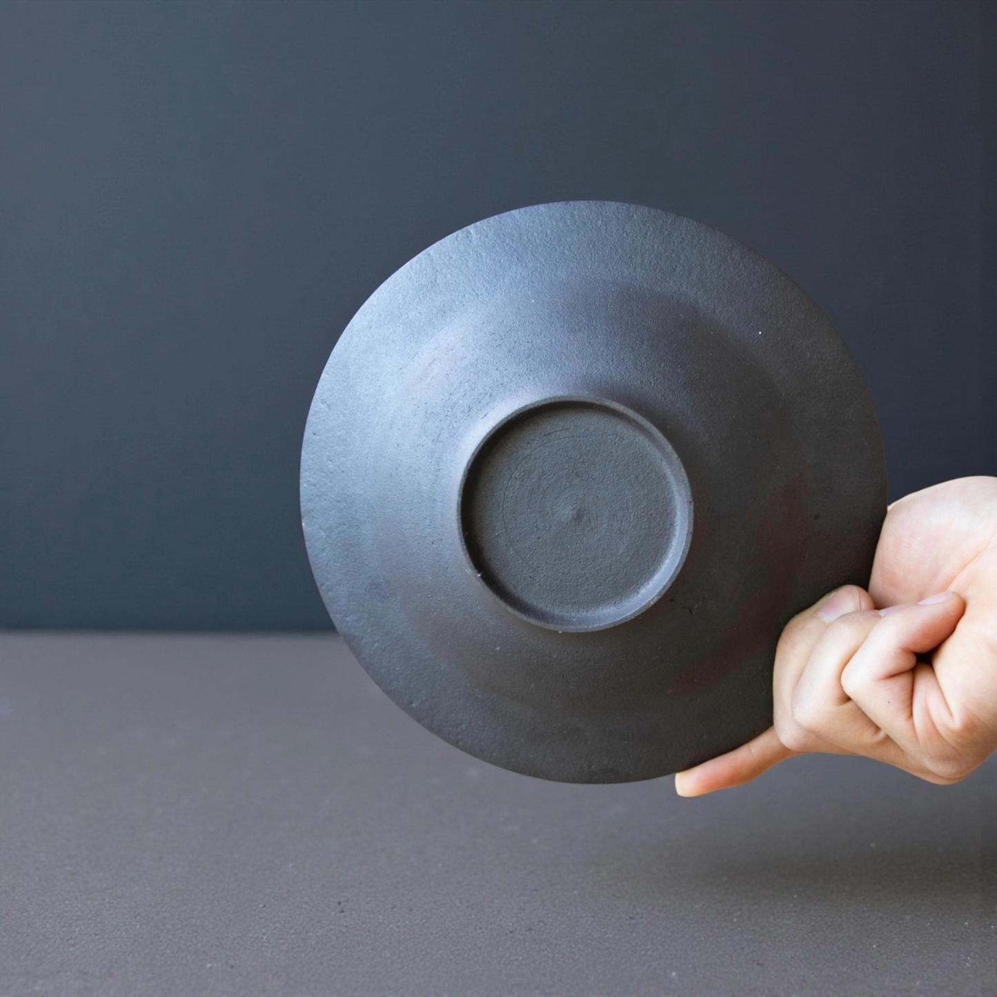 Hand holding a dark gray ceramic plate showing its bottom side against a dark gray background