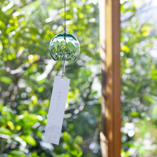 A green glass wind chime with a hanging white tag, photographed outdoors against a blurred background of green foliage and a wooden post.