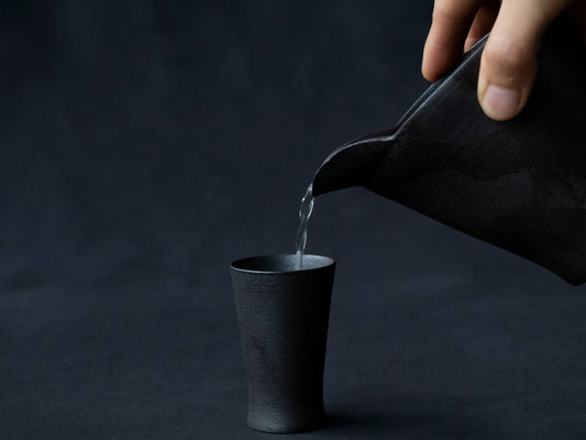 sake being poured into black ceramic cup  from black ceramic carafe by hand 